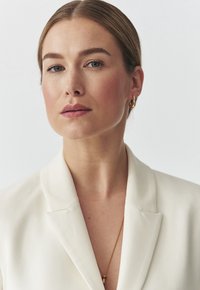 Bílý blejzr se strukturovaným límcem, nošený přes zlatý náhrdelník s přívěskem. Modelka má dlouhé vlasy a přirozený make-up.