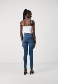 High-waisted blue denim jeans with a smooth texture, featuring two back pockets and a brown label at the waistband. Model wears a white top.