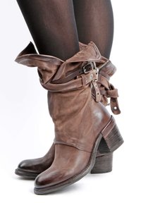Botas de cuero marrón hasta el tobillo con un puño doblado, correas decorativas y acentos metálicos. Tacón grueso de madera con una textura ligeramente desgastada.