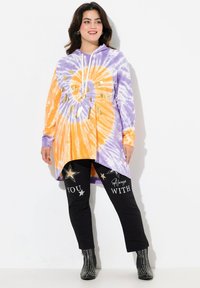 Tunika con cappuccio in tie-dye con motivi arancioni e viola, con accenti dorati. Indossata con pantaloni neri caratterizzati da testo bianco e stelle.