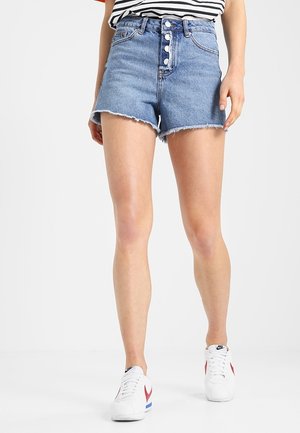 Vrouw draagt hooggetailleerde denim shorts met knoopsluiting en rafelige zomen, gecombineerd met witte Nike-sneakers en een gestreept shirt.