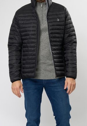 Mann trägt eine schwarze gesteppte Steppjacke über einem grauen Pullover mit halbhohem Reißverschluss und blauen Jeans, steht vor einem einfarbigen Hintergrund.