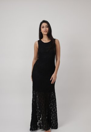 Femme debout dans une robe noire sans manches en dentelle avec une jupe transparente, sur un fond clair uni.