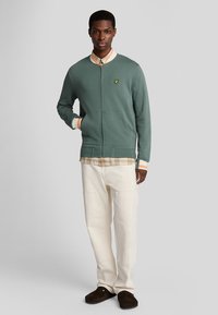 Sweat-shirt vert zippé avec des poignets et un ourlet côtelés, superposé sur une chemise à carreaux. Associé à un pantalon blanc et des chaussons marron.