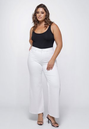 Woman with wavy brown hair wearing a black tank top, high-waisted white wide-leg pants, and black heeled sandals against a plain background.