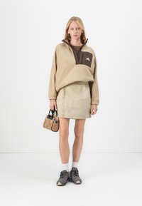 Beige fleecetrui met een bruine accent, beige spijkerrok, stevige donkere sneakers en een kleine bruine handtas. Witte achtergrond.