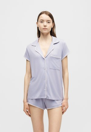Femme portant un haut de pyjama violet clair à manches courtes avec poche et short assorti, debout devant un fond blanc uni.