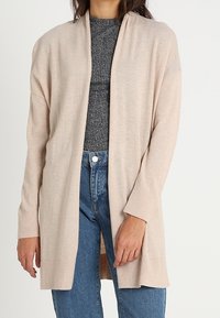 Mujer con un cárdigan beige de frente abierto sobre un suéter de canalé gris oscuro y jeans de talle alto azules, de pie contra un fondo neutro.