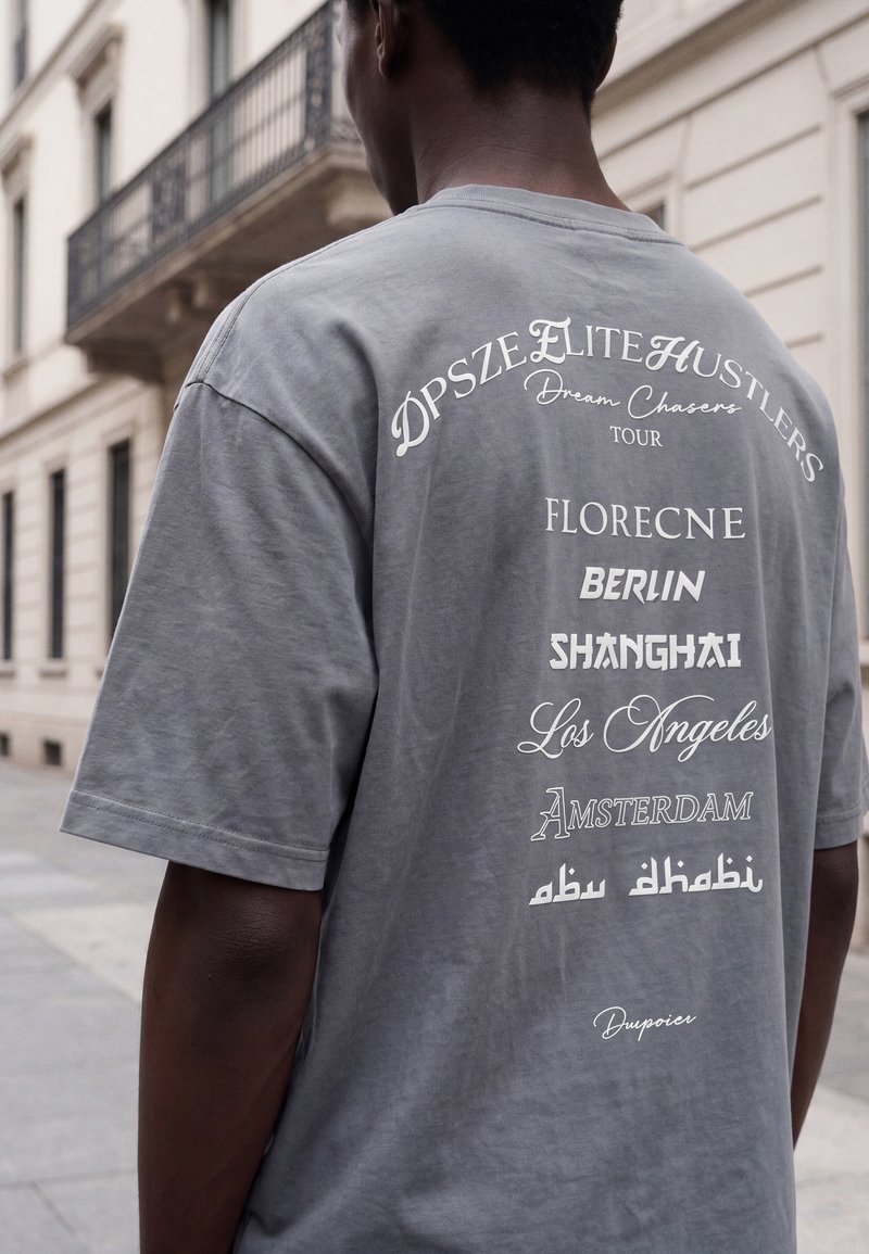 Persona che indossa una maglietta grigia con i nomi delle città del tour stampati in bianco sul retro, tra cui Berlino, Shanghai, Los Angeles e Abu Dhabi.
