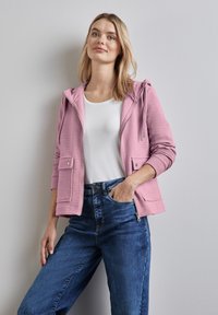 Light pink hooded jacket with a textured finish, featuring front pockets. Worn over a white top, paired with dark blue denim jeans.