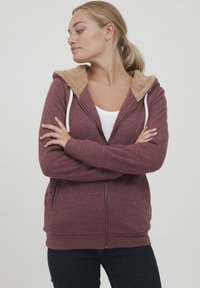 Bordo zip-up dukserica s kapuljačom obloženom bež plišem, rebrastim manžetama i opuštenim krojem. Ima vanjske džepove i zatvaranje na zip.