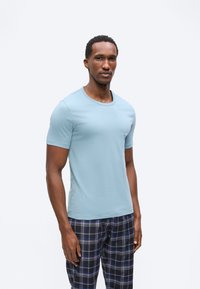 Άνδρας που φοράει ανοιχτό μπλε κοντομάνικη μπλούζα και μπλε και μαύρο καρό παντελόνι, στέκεται μπροστά σε απλό λευκό φόντο.