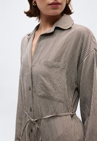 Γυναίκα φορώντας φόρεμα με κάθετες καφέ και λευκές ρίγες, κουμπωτό, με τσέπη στο στήθος και ζώνη στη μέση. Φοράει χρυσούς κρίκους σκουλαρίκια.