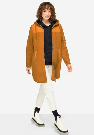 Orange fuzzy coat with a hood, contrasting smooth upper sections, zip front, side pockets, paired with a navy hooded top and white pants.