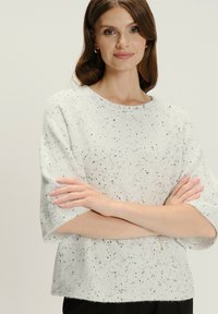 Kobieta z jasno-brązowymi włosami, ubrana w białą sweter w drobne plamki, z rękami skrzyżowanymi i delikatnym uśmiechem na tle jednolitym.