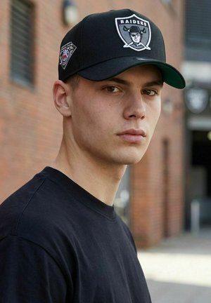 Junger Mann mit schwarzer Raiders-Kappe und schwarzem Hemd steht draußen vor einer Ziegelwand und blickt in die Kamera.