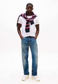 Vit t-shirt, blå jeans och vita sneakers. En marinblå och röd randig halsduk är hängd över axlarna. Enkel design och avslappnad passform.