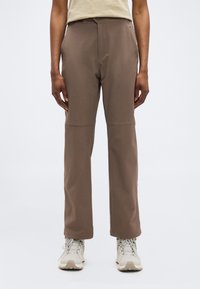 Άτομο που φορά καφέ παντελόνι ίσιας γραμμής και μπεζ παπούτσια πεζοπορίας, στέκεται μπροστά σε απλό λευκό φόντο.