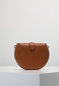 Braune Ledertasche mit Crossbody-Design und runder Form. Verfügt über einen Klappenverschluss und dezente Nähte, präsentiert auf einem weißen Podest.