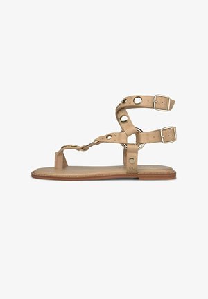 Flache Sandale aus hellbraunem Leder mit Knöchelriemen, goldfarbenen runden Nieten und seitlichen Schnallen auf einer hellbraunen Sohle.