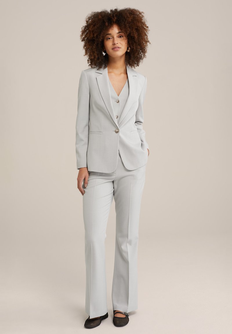Costume sur mesure gris clair avec un blazer à un bouton, des revers pointus et un pantalon droit. Tissu texturé avec un léger éclat.
