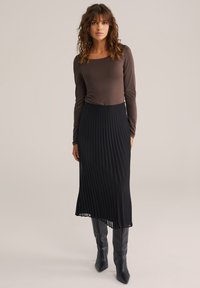 Ruda ilgomis rankovėmis palaidinė derinama su juoda plisuočiuota midi sijonu. Modelis vilki aukštus juodus batelius su smailiu nosiu, pristatydama elegantišką siluetą.