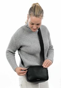Borsa a tracolla nera in pelle con finitura testurizzata, forma rettangolare, chiusura con zip e tracolla regolabile, indossata davanti a un maglione grigio.
