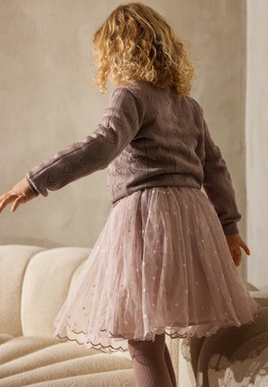 Kind met krullend blond haar dat een paarse trui met hartjespatroon en een roze tulen rok draagt, staand met gespreide armen op een beige bank.