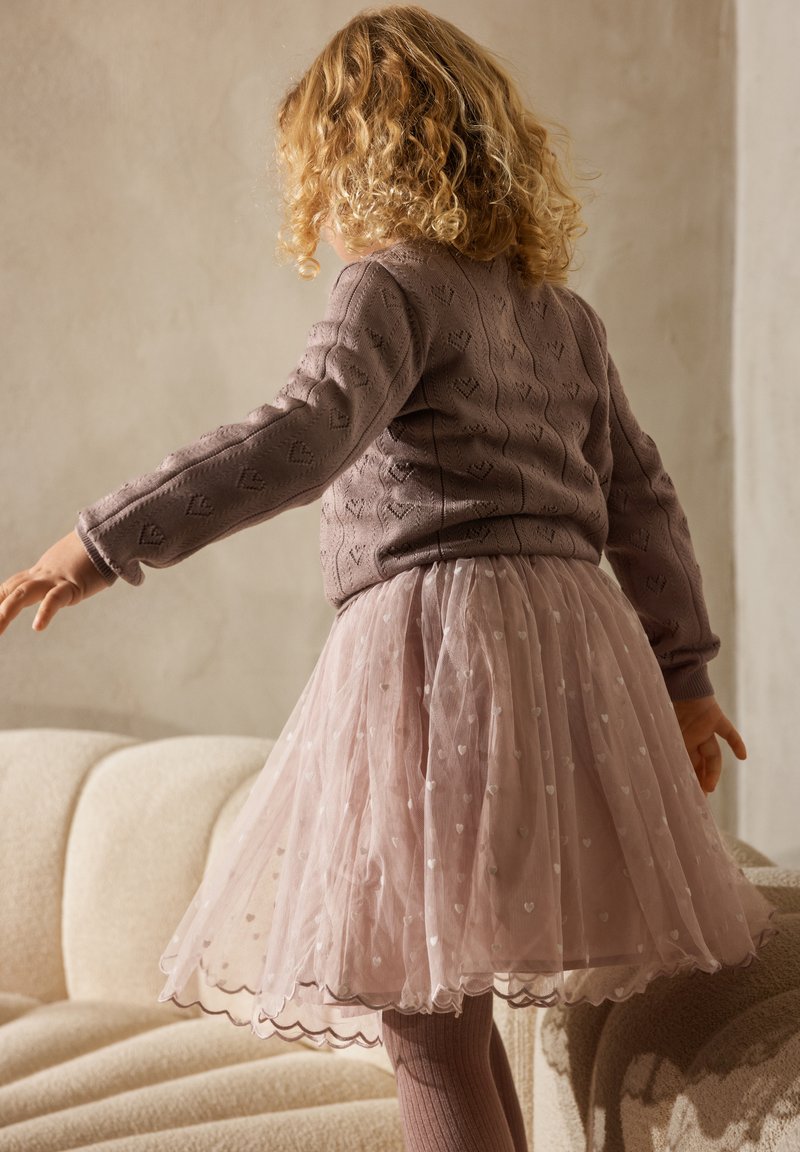 Enfant aux cheveux blonds bouclés portant un pull violet à motifs de cœurs et une jupe en tulle rose, debout les bras écartés sur un canapé beige.