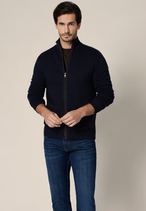 Mężczyzna ubrany w ciemnogranatowy sweter zapinany na zamek, na czarnej koszulce i niebieskich dżinsach, stoi na tle jednolitego beżowego tła.