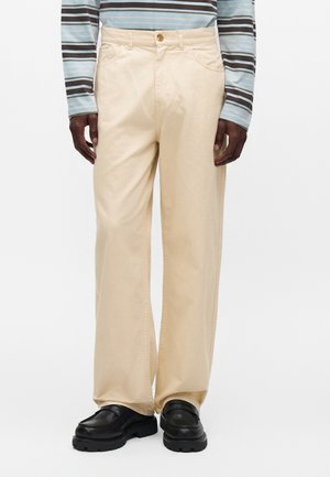 Person iført beige bukser med vide ben, en lyseblå og brunstribet langærmet skjorte og sorte chunky loafers, står mod en ensfarvet baggrund.