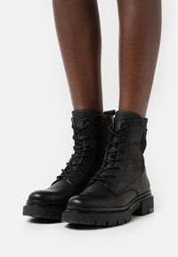 Botas de cuero negro con cordones en la parte delantera, lados texturizados y suela resistente. Incluyen una cremallera y un collar acolchado para mayor comodidad.