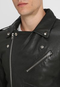 Veste de moto en cuir noir avec un large col, des accessoires en métal argenté, des poches zippées et une texture lisse.