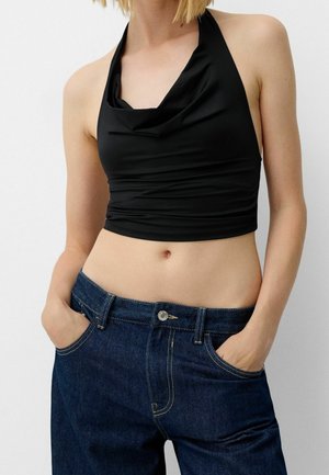 Kvinde iført sort halterneck croptop og mørkeblå højtaljede jeans med hænderne i lommerne mod en ensfarvet baggrund.