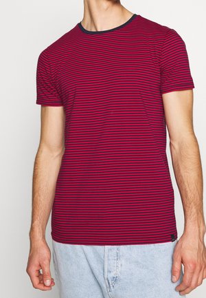 Homme portant un t-shirt à manches courtes à fines rayures rouges et bleu marine, ainsi qu'un jean en denim bleu clair, debout devant un fond uni.