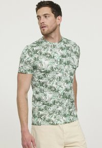 T-shirt en imprim é botanique vert, fabriqué en tissu douce, avec des manches courtes et un col rond. Le motif comprend diverses feuilles vertes sur un fond blanc.