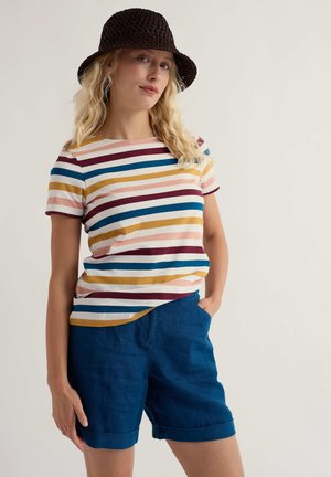 Nő fekete kötött horgászkalapban, színes csíkos pólóban és kék, felhajtott szegélyű rövidnadrágban, egyik kezét a zsebébe dugva áll.