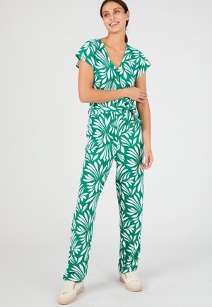 Vrouw draagt een groen-wit jumpsuit met bladmotief en korte mouwen, staat met één arm gekruist en lacht.