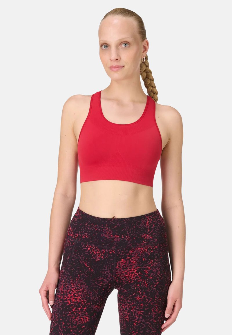 Femme aux cheveux tressés portant un brassière de sport rouge et un legging noir avec des motifs abstraits rouges, debout devant un fond clair uni.