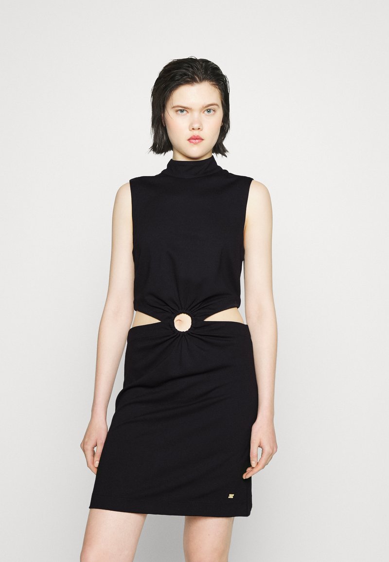 Vestido preto sem mangas com um decote alto, apresentando um detalhe em nó e recorte na cintura, feito de tecido suave, com um detalhe dourado.