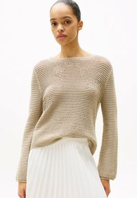 Beige gestrickter Pullover mit lockerer Passform, weiter Ausschnitt und langen Ärmeln, kombiniert mit einem weißen, plissierten Rock. Die Textur ist offen gewebt.