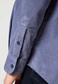 Camisa de pana abotonada en azul oscuro con textura acanalada, que presenta un detalle de puño en ángulo y un solo botón azul en la manga.