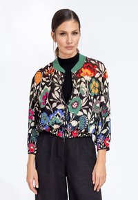 Giacca bomber con zip a fantasia floreale su base nera, caratterizzata da fiori multicolori e colletto a costine verde, abbinata a pantaloni neri.