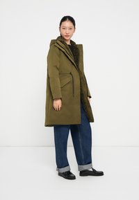 Parka verde oliva con cappuccio, dotata di imbottitura soffice, due tasche frontali e cordoncino in vita. Indossata sopra un paio di jeans blu.