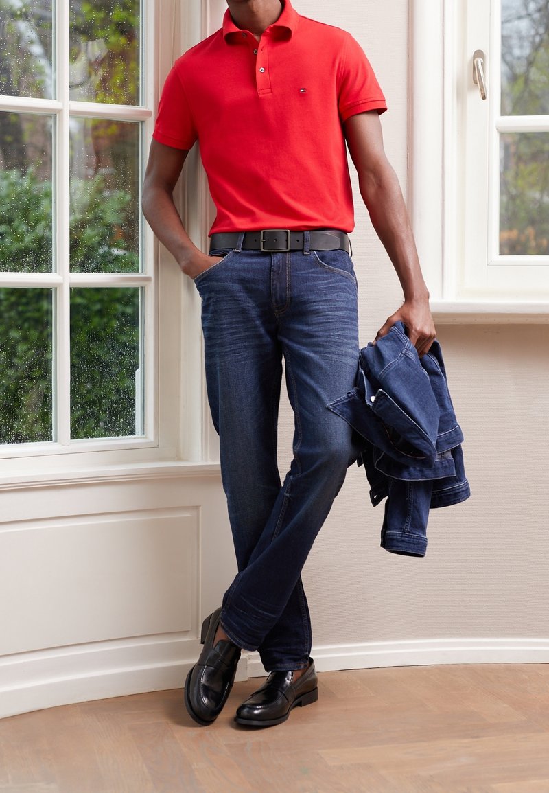 Homme portant un polo rouge, un jean bleu et des chaussures noires, debout à l'intérieur près des fenêtres, tenant une veste en jean bleue pliée dans une main.
