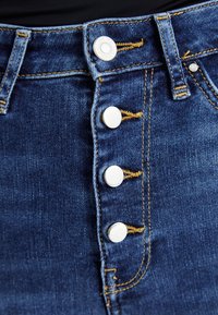 Jupe en denim avec quatre boutons-pression argentés brillants, des coutures jaunes et une taille ajustée, de couleur bleu foncé et avec une texture lisse.