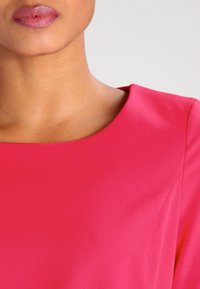 Fuchsiové růžové šaty s hladkou texturou, kulatým výstřihem a čistými lemy, které zdůrazňují přiléhavý střih. Žádné viditelné vzory ani akcenty.