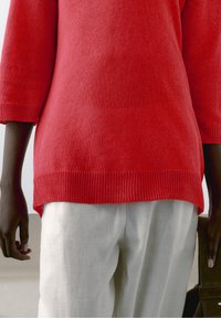 Czerwony sweter z dzianiny z rękawami do połowy łokcia i prążkowanym dołem, zestawiony z jasnymi lnianymi spodniami. Gładka faktura, luźny krój.