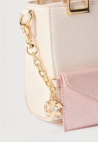 Sac à main en texture crème avec une pochette enveloppe rose attachée par une chaîne dorée ornée d'un charme en forme de fleur et de fer à cheval.