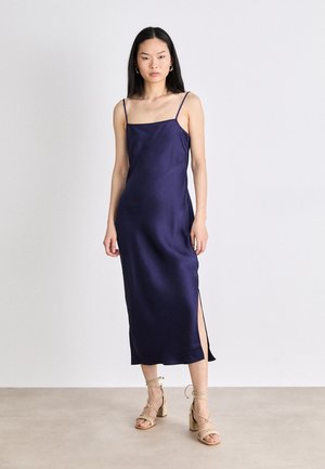 Robe satin bleu marine avec des bretelles fines, fente sur le côté et ourlet droit. Associée à des sandales à talons à lanières de couleur beige clair.